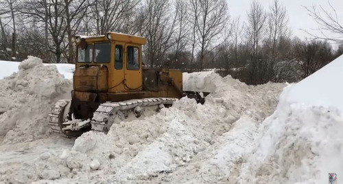Водитель бульдозера расчищает дорогу после снегопада. Стопкадр из видео https://www.youtube.com/watch?v=Fe9qu-qUVH8