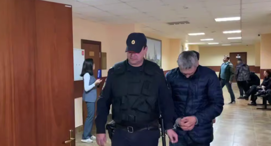 Конвоир ведет одного из подсудимых. Стоп-кадр видео "Фортанга" от 20.05.25, https://t.me/fortangaorg/19717?single