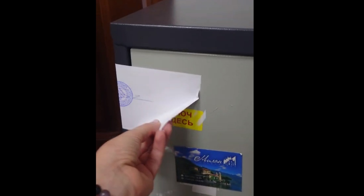 Автор видео демонстрирует отклеенный угол листа бумаги, которым опечатан сейф. Кадр видео телеграм-канала "ТИК Западная голосование 2026" https://t.me/tikzapad/91
